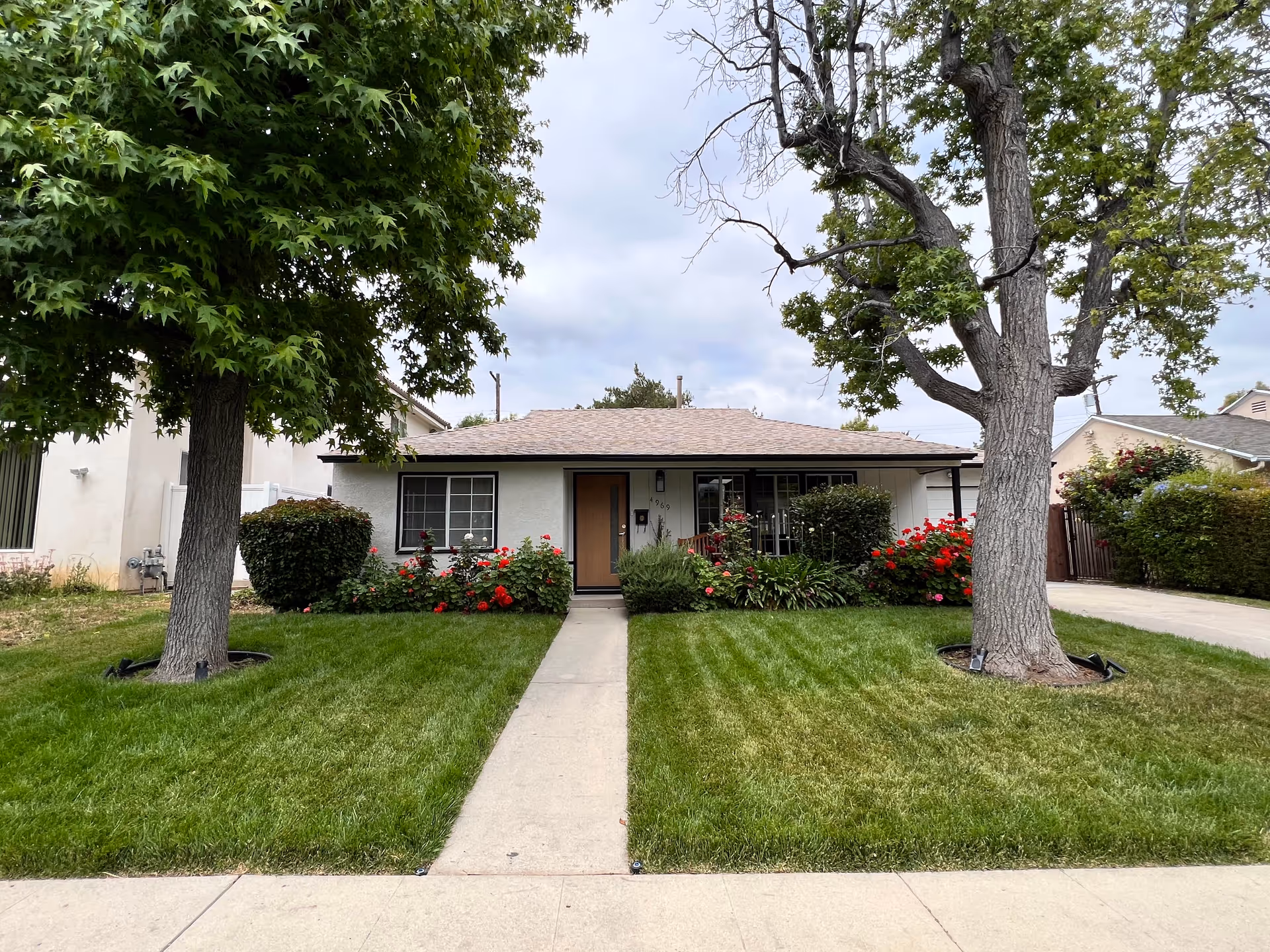 Single-story house front with a central walkway, green lawn, two large trees, and flower beds.
