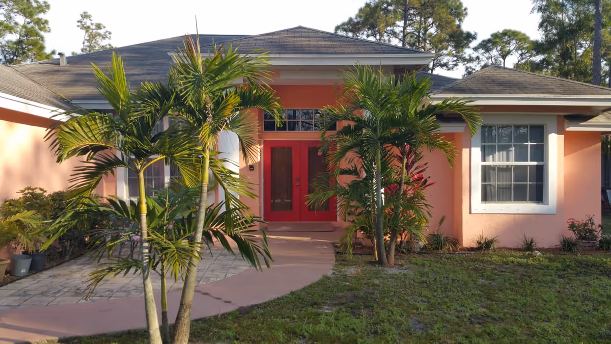 Pink single-story building front with a red door, palm trees, and a small lawn.
