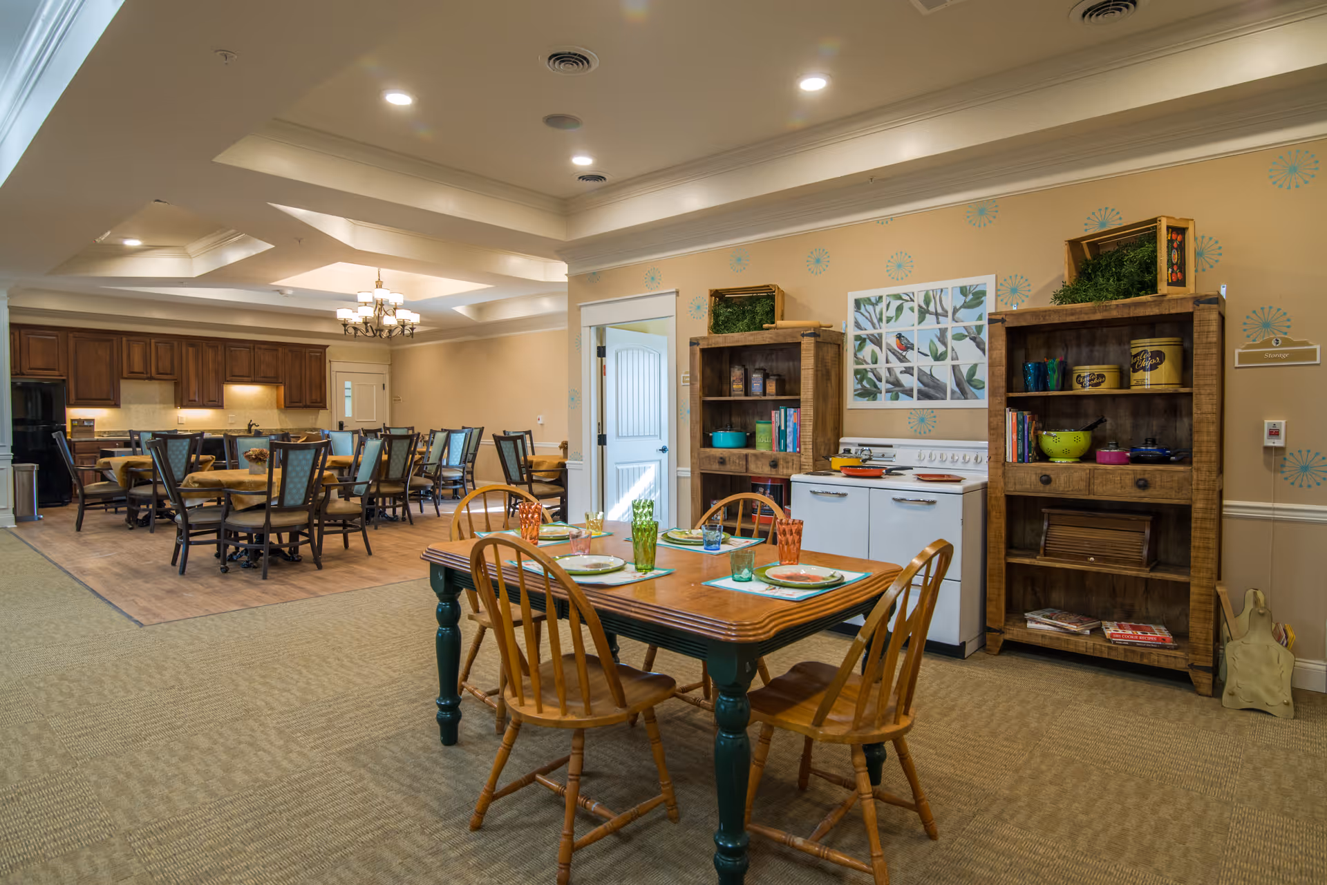 A dining area in a senior living facility featuring a wooden table set for four with colorful glasses and plates. Behind the table are two wooden shelves filled with decorative items and kitchenware, and a white stove with a bird-themed artwork above it. In the background, there are multiple dining tables and chairs, a kitchen area with wooden cabinets, and a chandelier hanging from the ceiling.