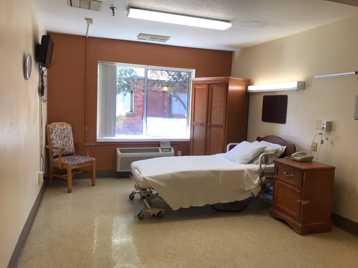 A simple, clean patient room with a hospital bed covered in white sheets, a wooden nightstand with a telephone, a wooden wardrobe, a floral upholstered chair, a wall-mounted TV, and a large window letting in natural light.