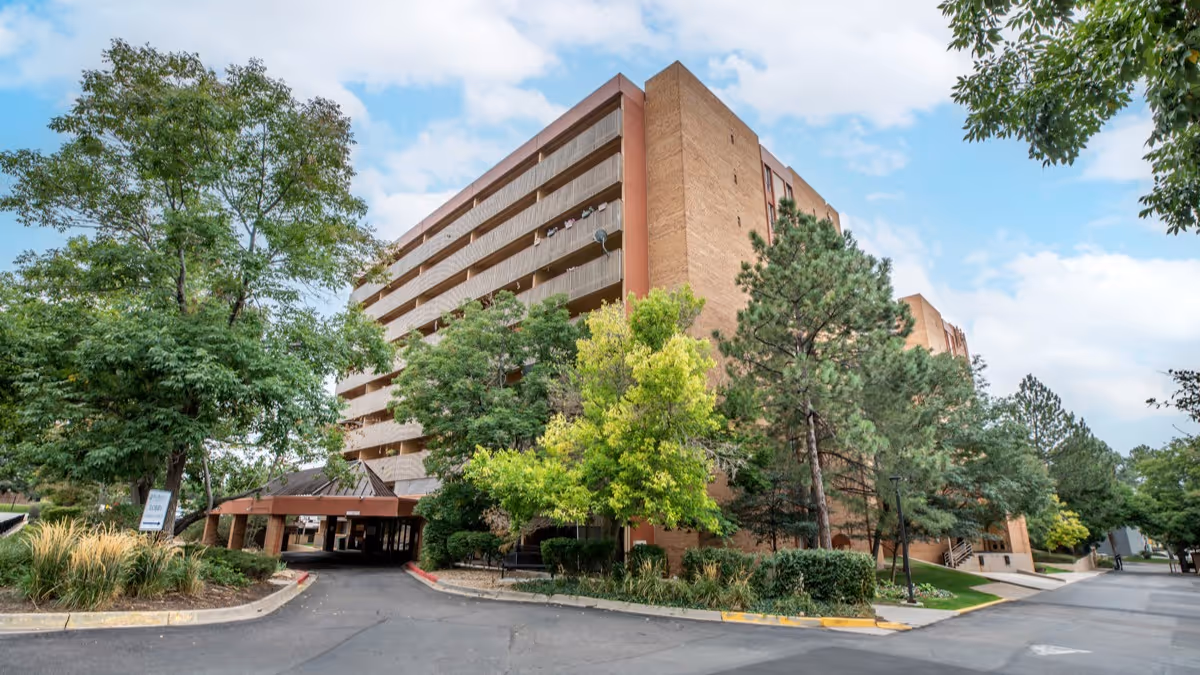 Exterior view of a multi-story senior living facility building surrounded by trees and greenery under a partly cloudy sky. The building has balconies and a covered entrance driveway.