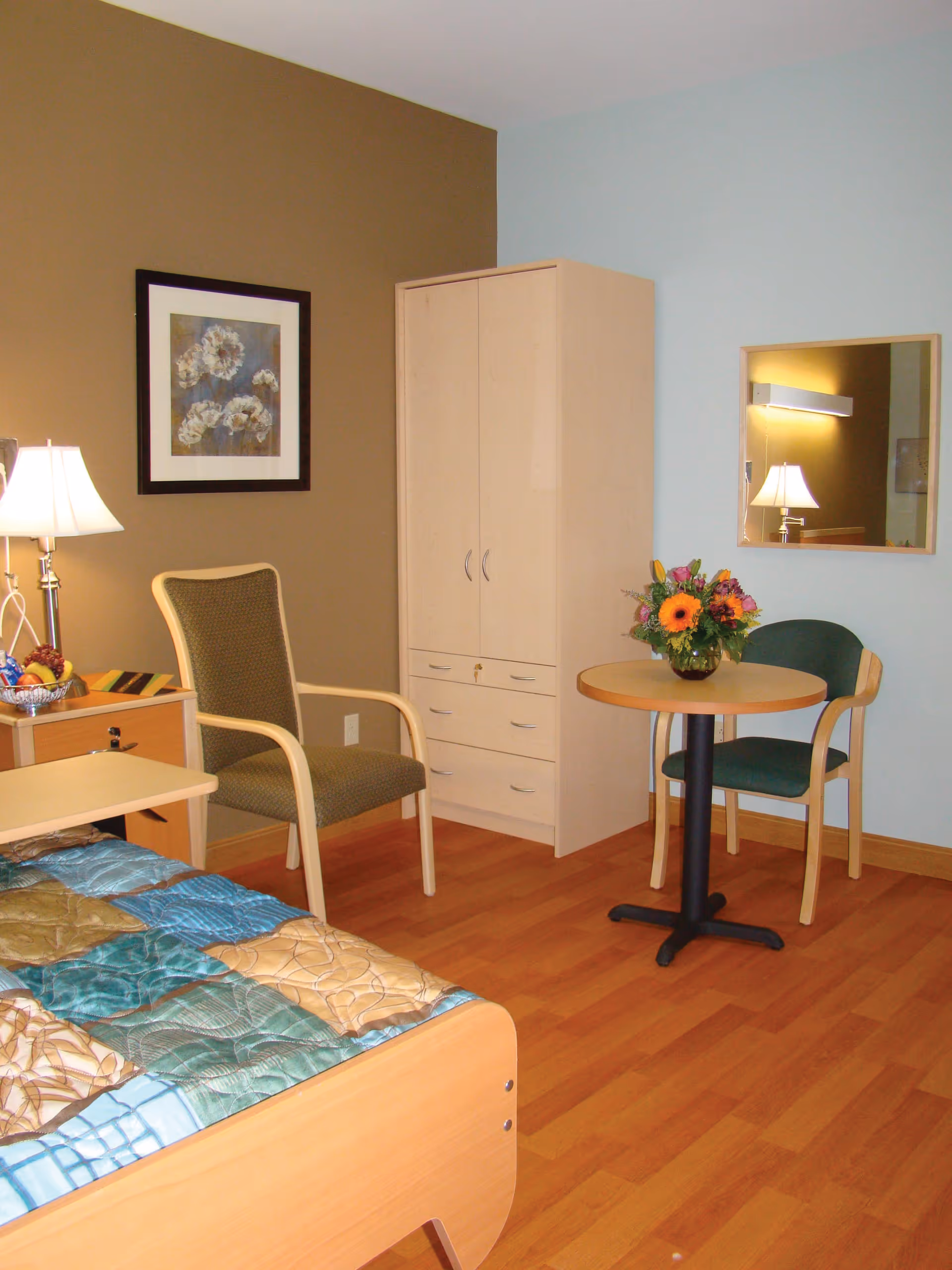 A cozy bedroom with a wooden bed covered by a multicolored quilt, a green cushioned chair, a small round table with a flower arrangement, a wooden wardrobe, a framed floral picture on the wall, and a lamp on a bedside table.