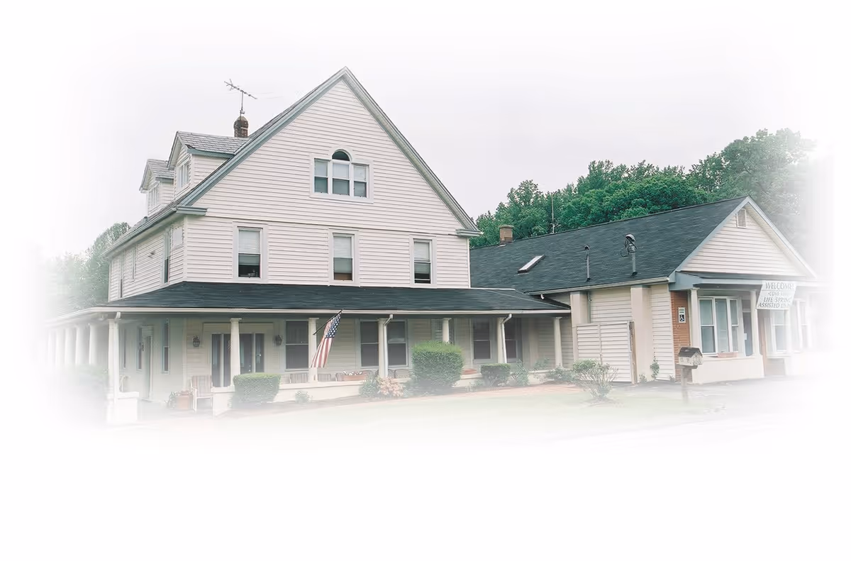 Two-story beige assisted living building with a wraparound porch and American flag.