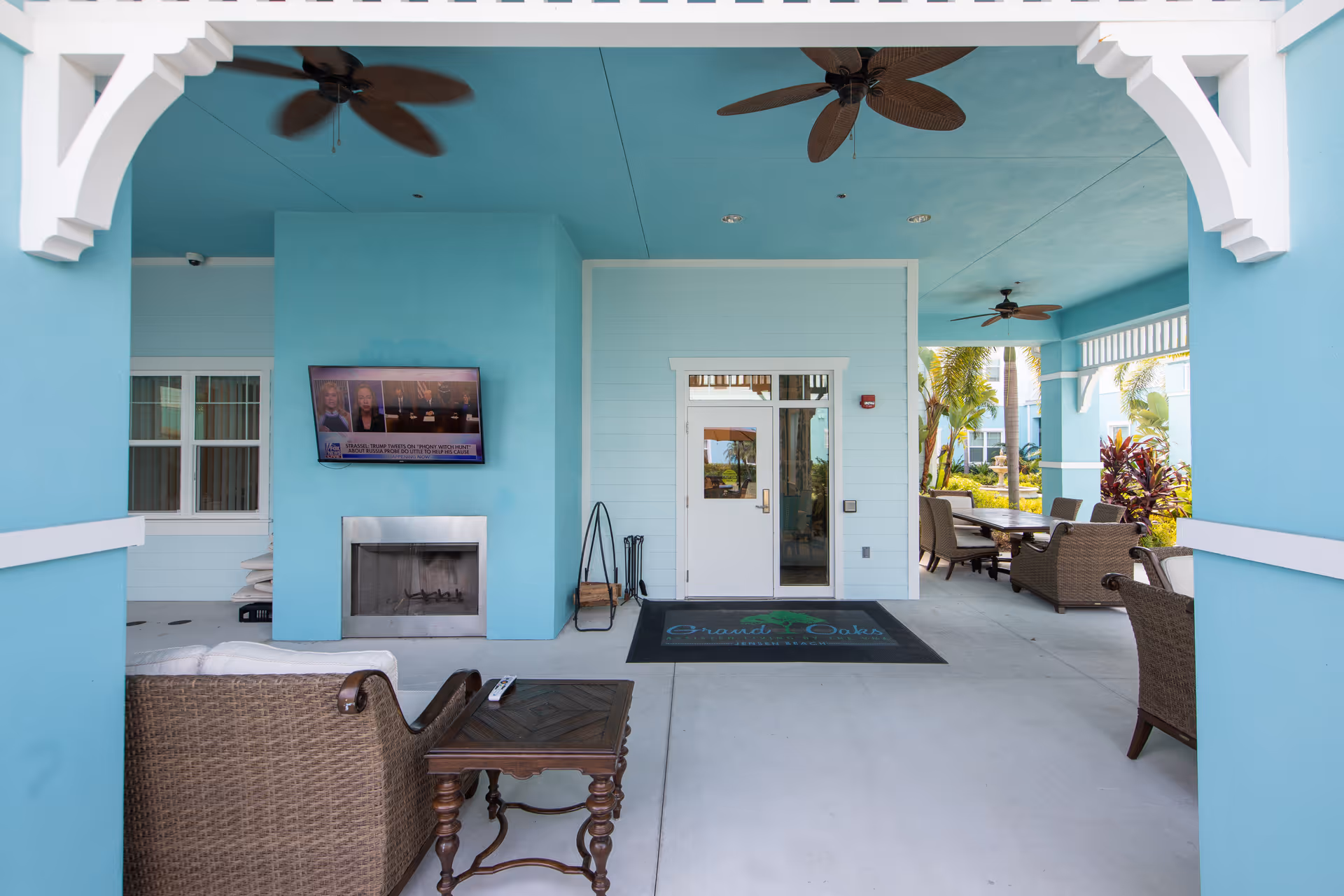 Covered turquoise entrance patio with wicker seating, wall-mounted TV and fireplace facing the glass entry doors.