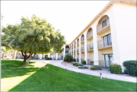 Exterior view of a three-story assisted living building with balconies, a paved walkway, green lawn, and a large tree.