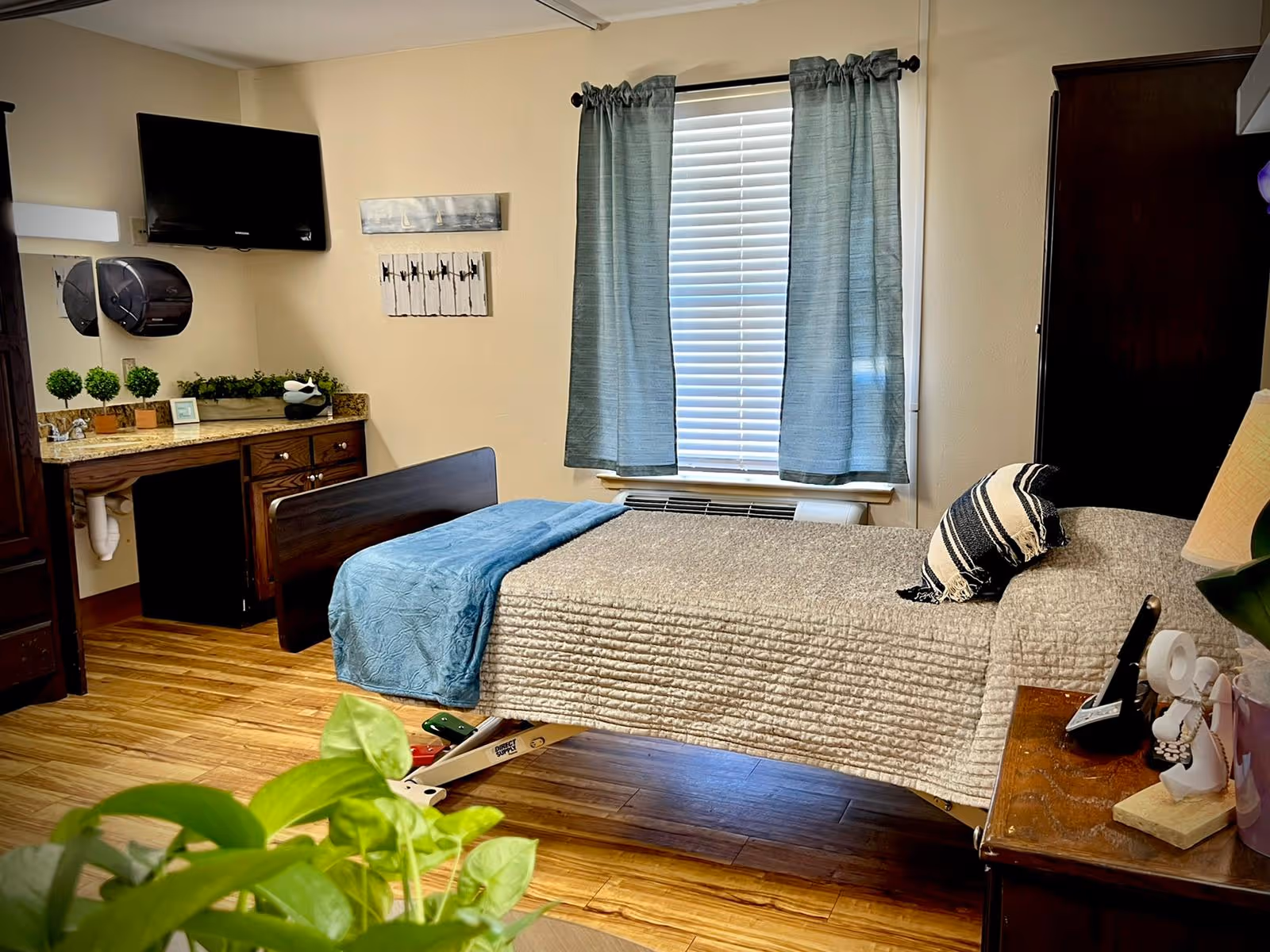A cozy bedroom in a senior living facility with a single bed covered with a beige quilt and a blue throw blanket. There is a striped pillow on the bed. The room has wooden flooring, a window with blue curtains, a wall-mounted TV, a wooden dresser with a granite countertop, and small decorative plants. A bedside table holds a lamp and some personal items. A green plant is partially visible in the foreground.