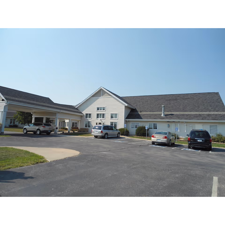 Front exterior of a single-story care facility with a covered drop-off, parked cars, and a paved parking lot.