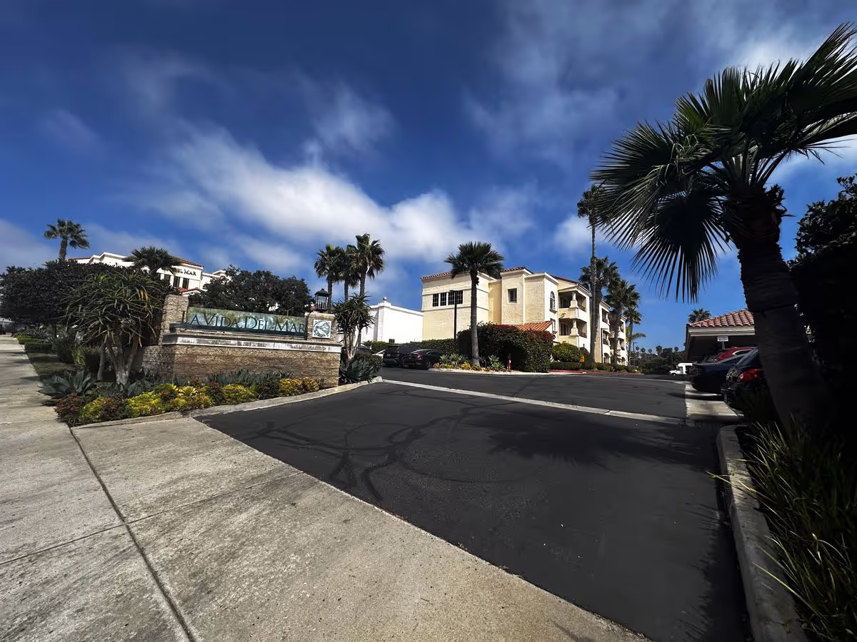 Entrance sign for La Vida Del Mar with a Mediterranean-style building, palm trees, driveway and blue sky.