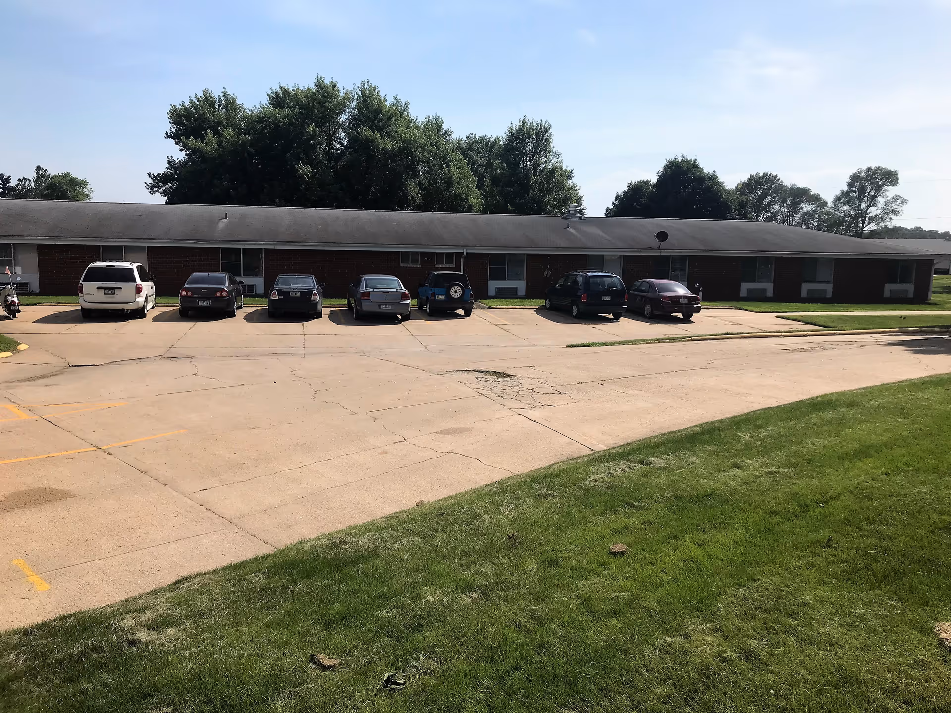 Long, single-story brick facility with a front parking lot, green lawn, and trees.