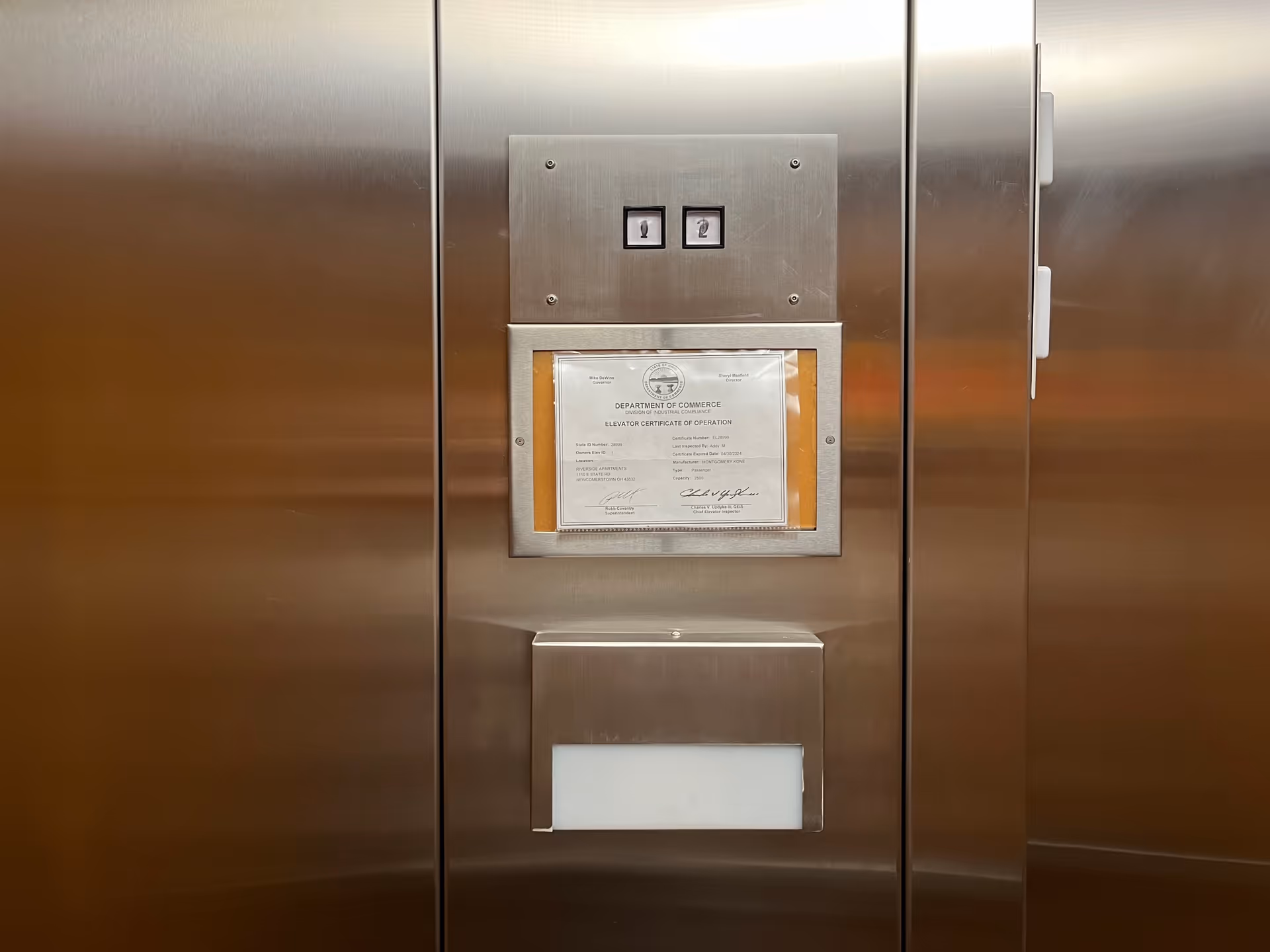 Stainless steel elevator interior with a central control panel and framed certificate.
