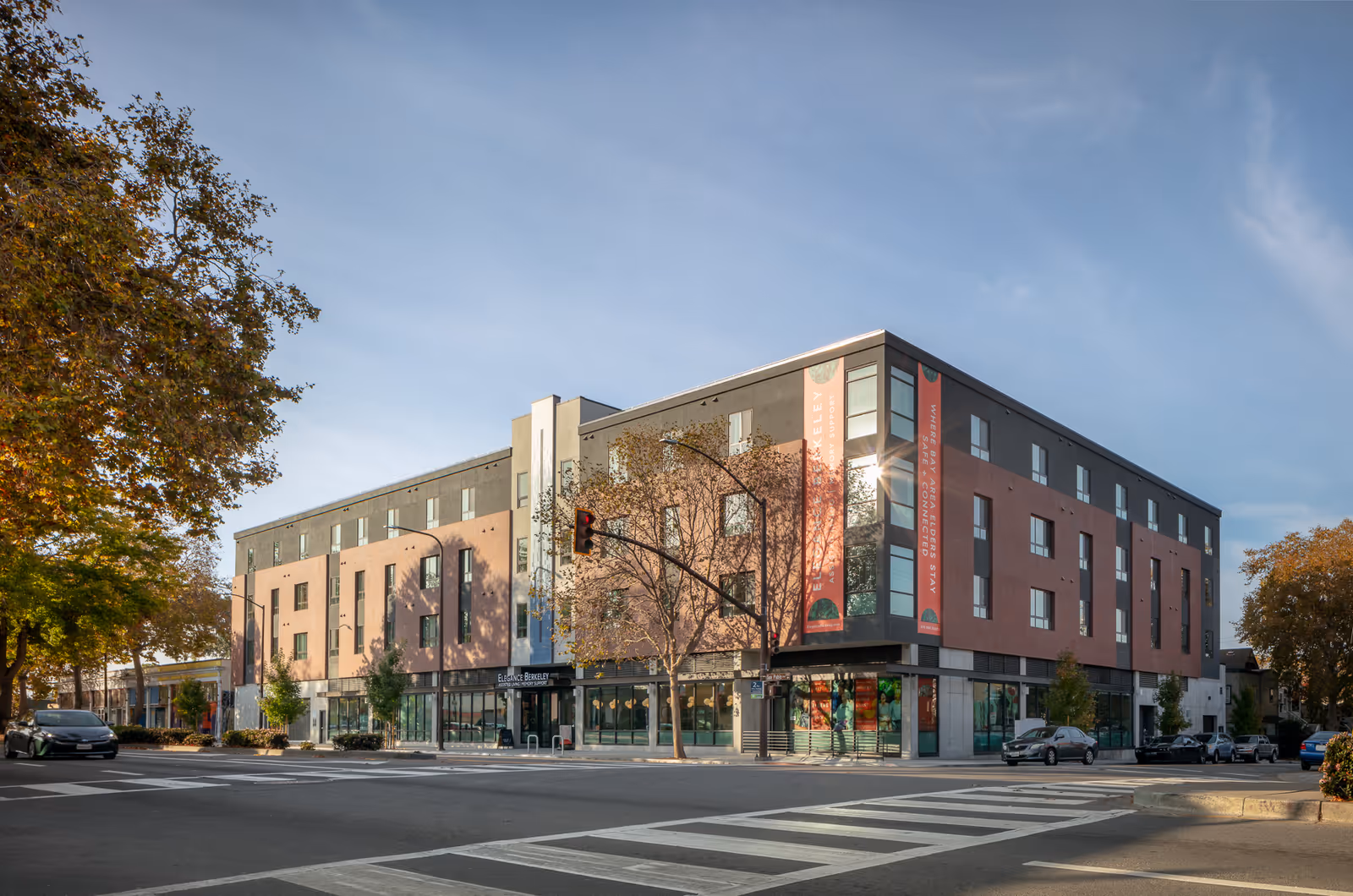 Exterior view of a modern four-story building named Elegance Berkeley located at a street corner with trees and parked cars nearby under a clear sky.