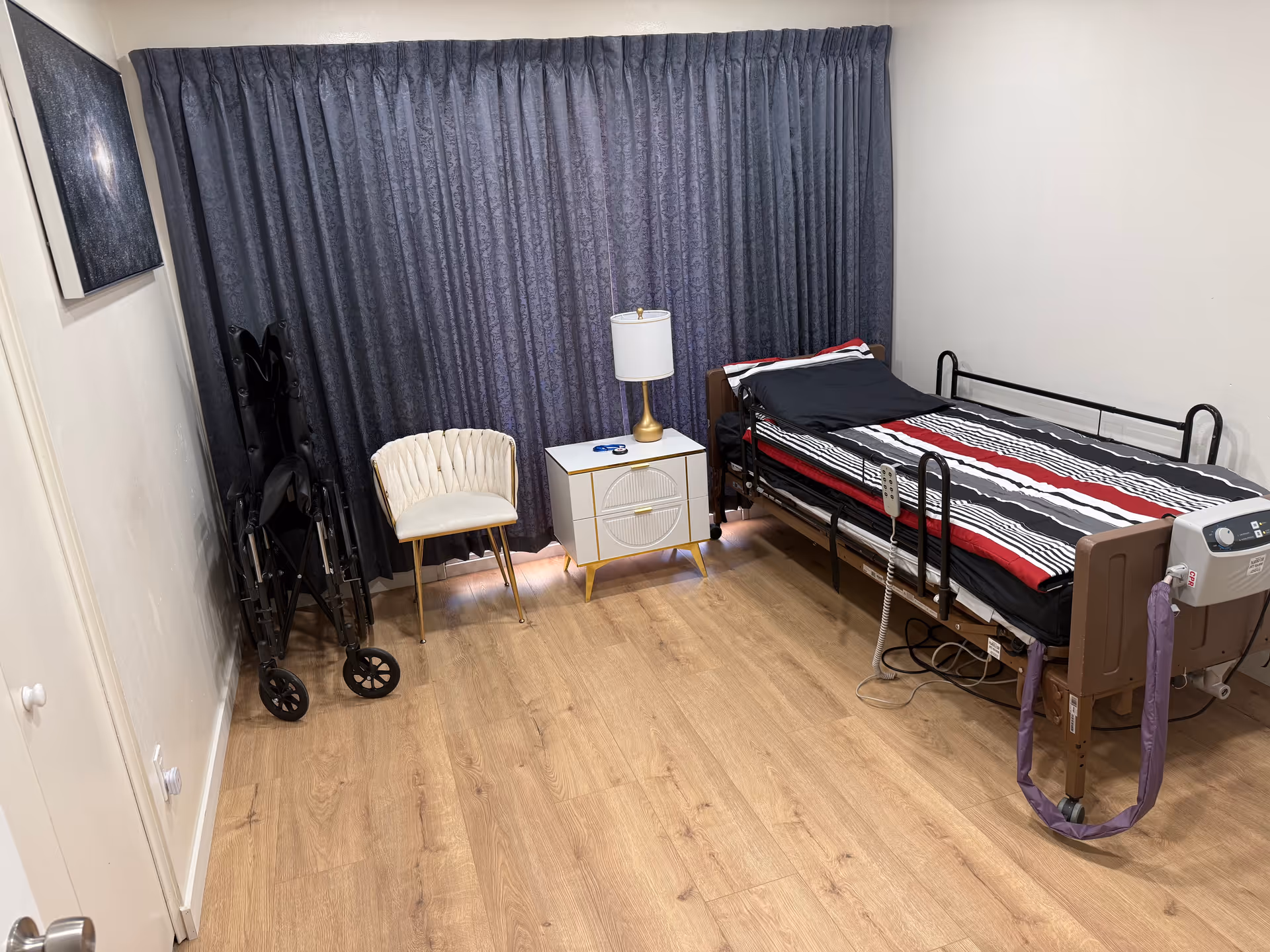 Small furnished bedroom with a hospital bed, nightstand, lamp, curtain backdrop, and mobility aid.