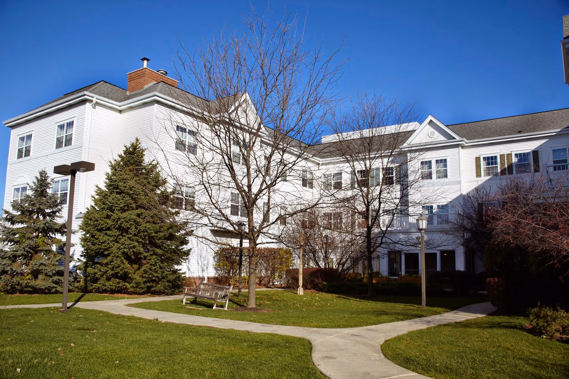 White three-story senior living building with leafless trees, benches, and a curved sidewalk across a green lawn under a clear blue sky.