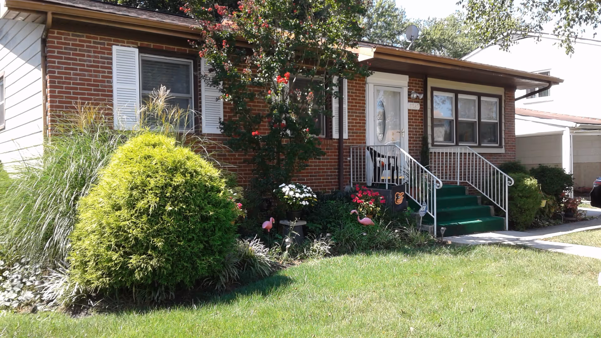 Front exterior view of a single-story brick house with white shutters and a white front door. There are green bushes, flowering plants, and two pink flamingo lawn ornaments in the garden. A green carpet covers the steps leading to the entrance, which has white railings on both sides.