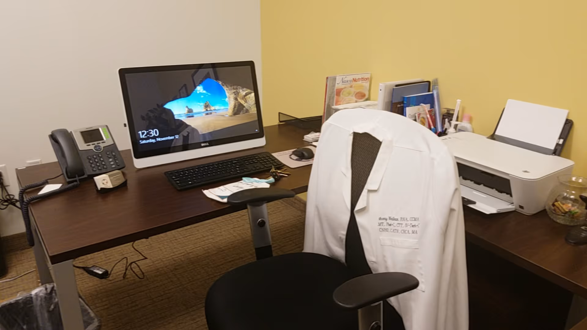Office desk with computer, phone, printer, and a white lab coat over a chair.