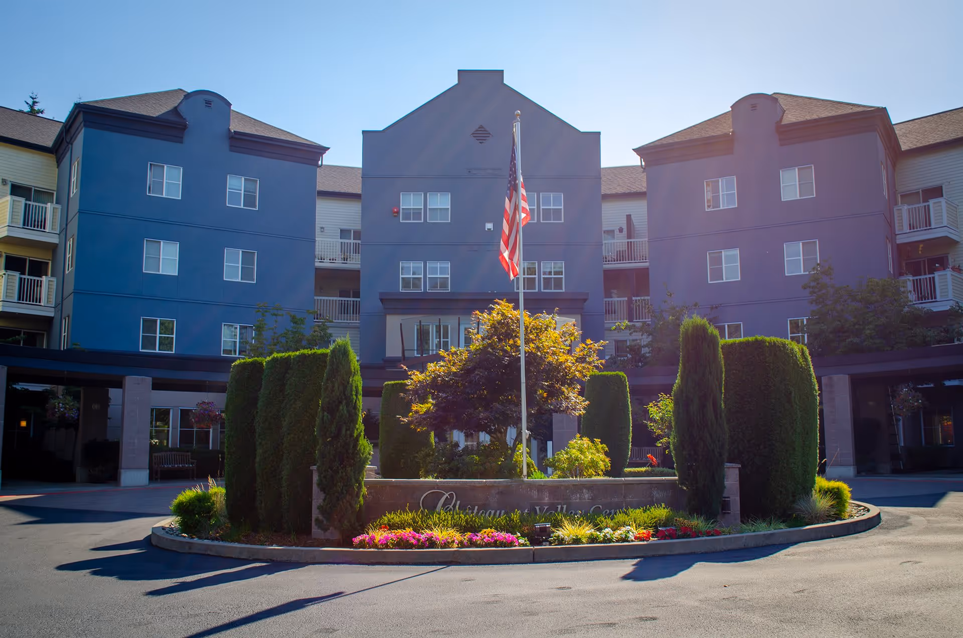 Front exterior view of a multi-story retirement facility building with a central landscaped area featuring trimmed bushes, flowers, and an American flag on a flagpole. The building has multiple windows and balconies under a clear blue sky.