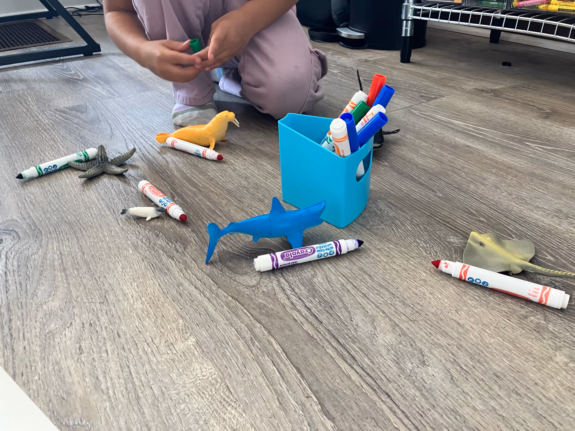 Child kneels on a wooden floor crafting with colorful glue, markers, and toy sea creatures.