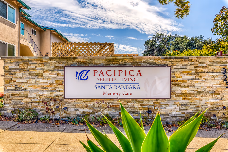 Stone wall with a sign that reads 'Pacifica Senior Living Santa Barbara Memory Care' in front of a residential building under a partly cloudy sky with greenery and a large plant in the foreground.