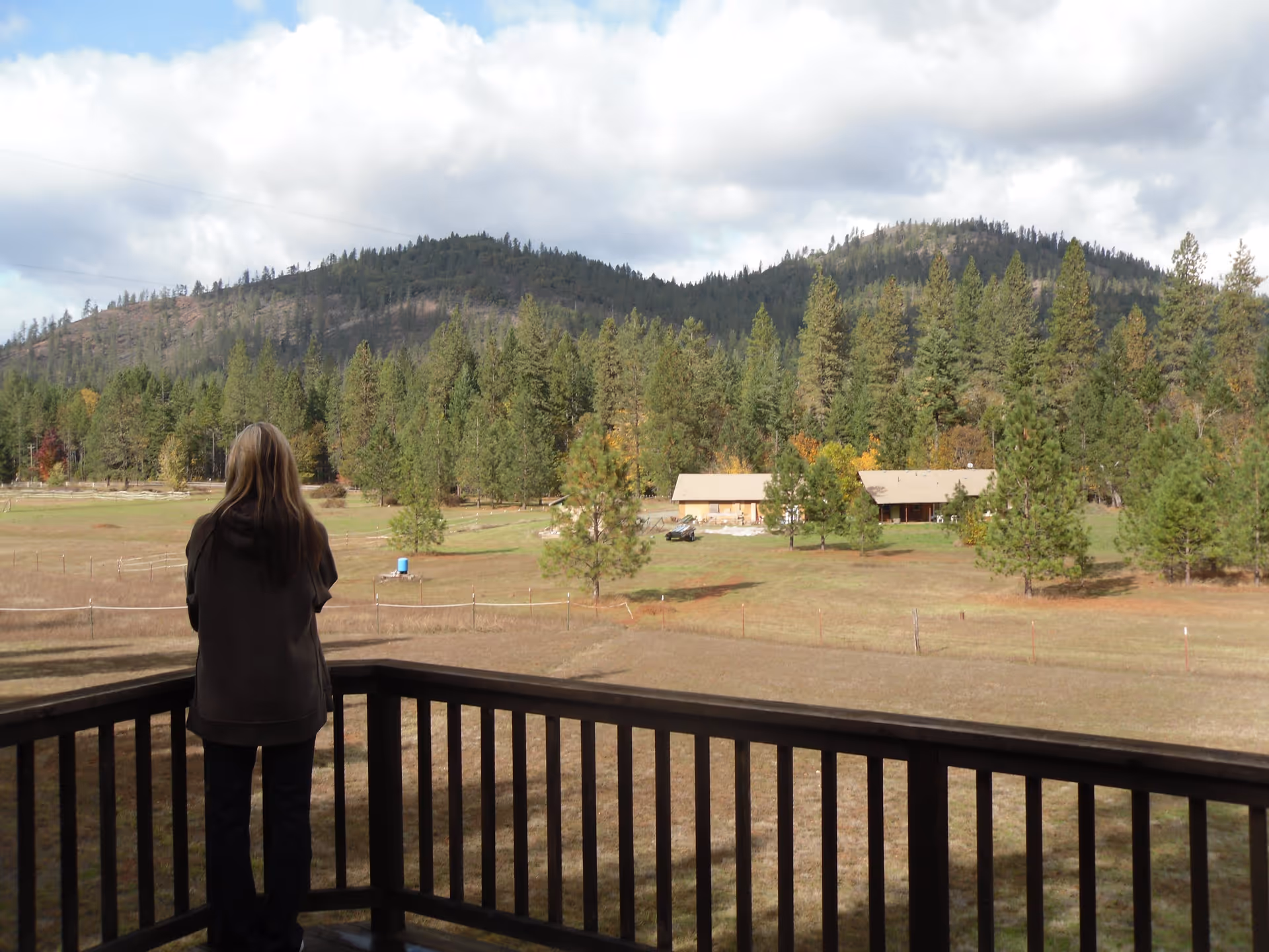 A person with long hair standing on a wooden deck railing, looking out over a large open field with scattered trees and two buildings in the distance, surrounded by forested hills under a partly cloudy sky.