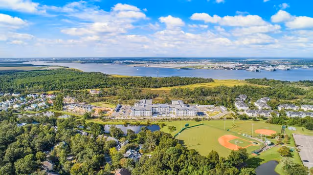 Aerial view of Overture Daniel Island senior living facility surrounded by greenery, a baseball field, and nearby water body under a partly cloudy blue sky.