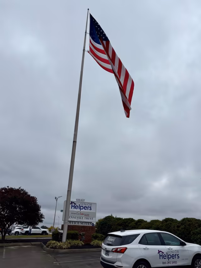 American flag on a tall pole beside a Senior Helpers sign and a parked van.