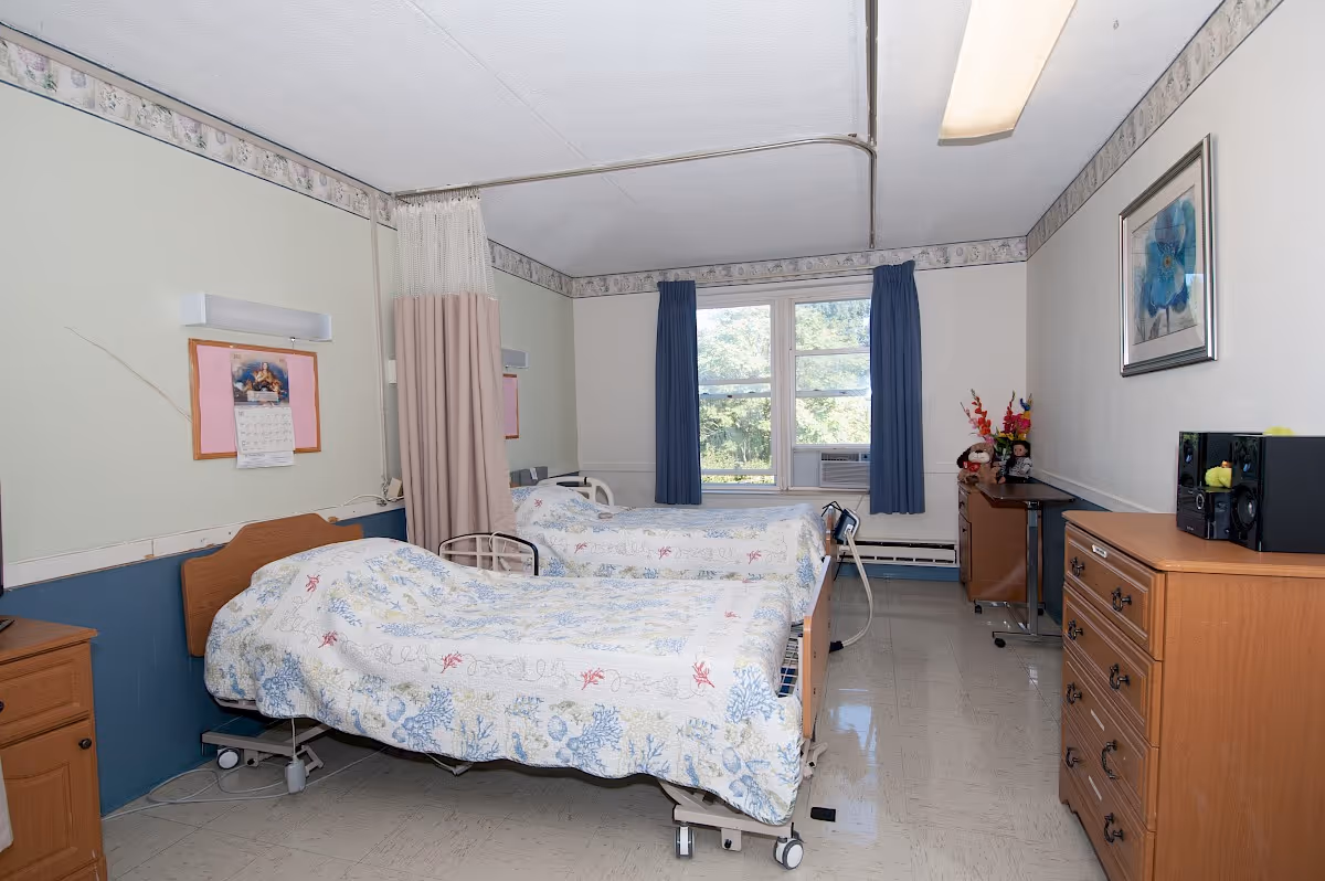 A shared bedroom in a care facility with two hospital-style beds covered in floral patterned bedspreads. The room has a window with blue curtains, a wooden dresser, a small table with flowers and decorations, and a privacy curtain between the beds. The walls are light-colored with a floral border near the ceiling.