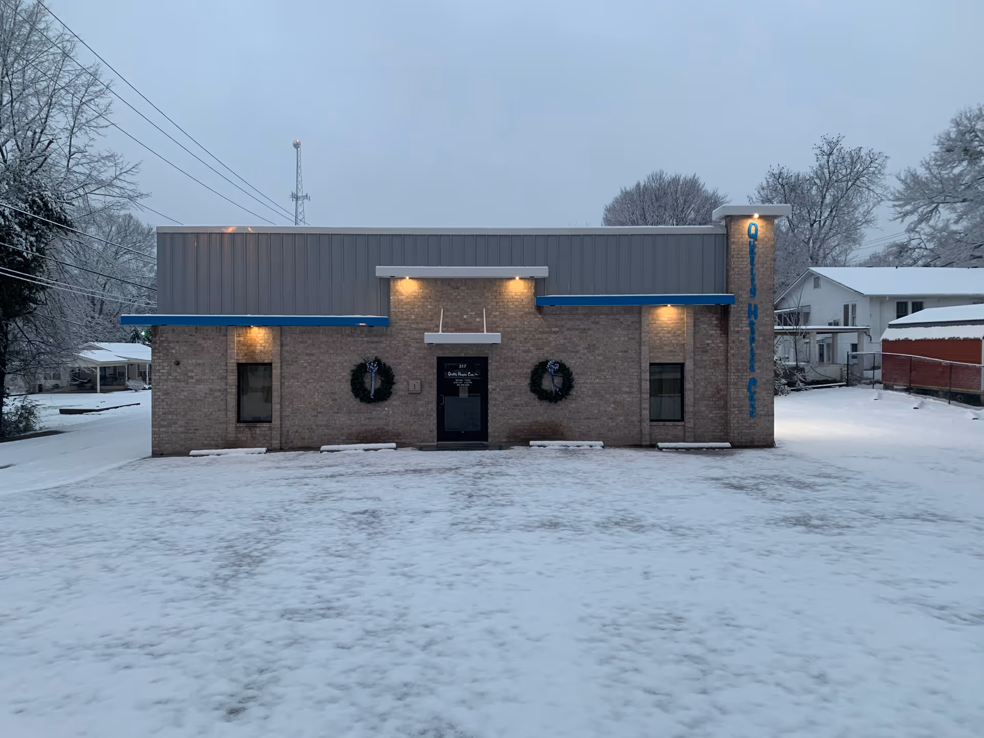 Snowy front of a brick building with blue awnings and wreaths, labeled Quality Hospice Care.