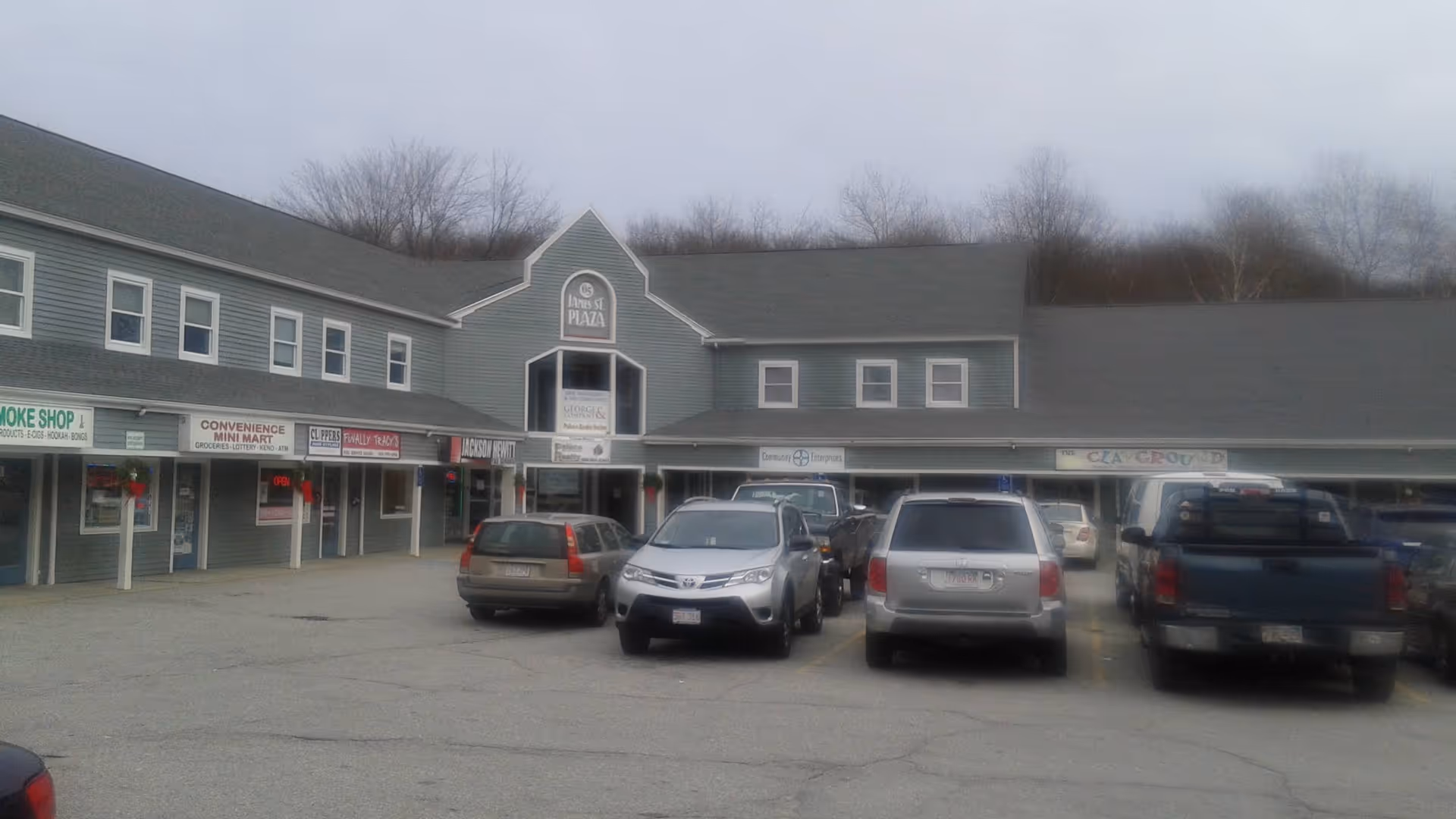 Row of storefronts in a strip mall with parked cars in front.