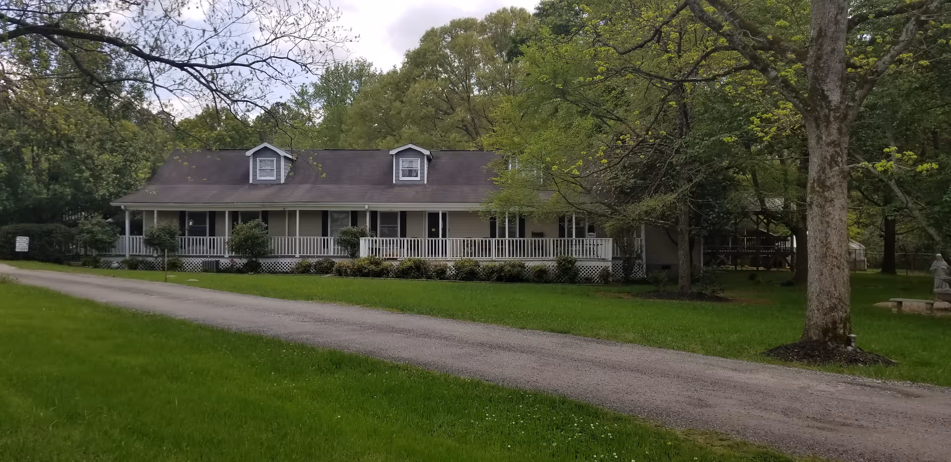 A single-story house with a long front porch and white railing, surrounded by green grass and trees. A gravel driveway leads up to the house, and there is a statue and bench visible on the right side near a large tree.