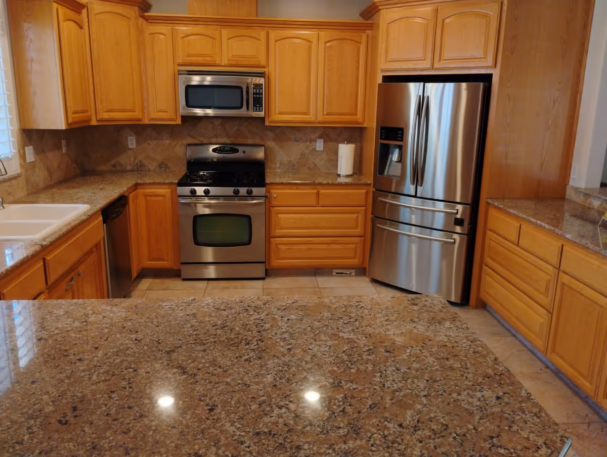 A kitchen with wooden cabinets, granite countertops, a stainless steel refrigerator with a water and ice dispenser, a stainless steel oven with a gas stove, a microwave above the stove, a dishwasher, and a white sink. The floor is tiled and the backsplash has a beige tile design.