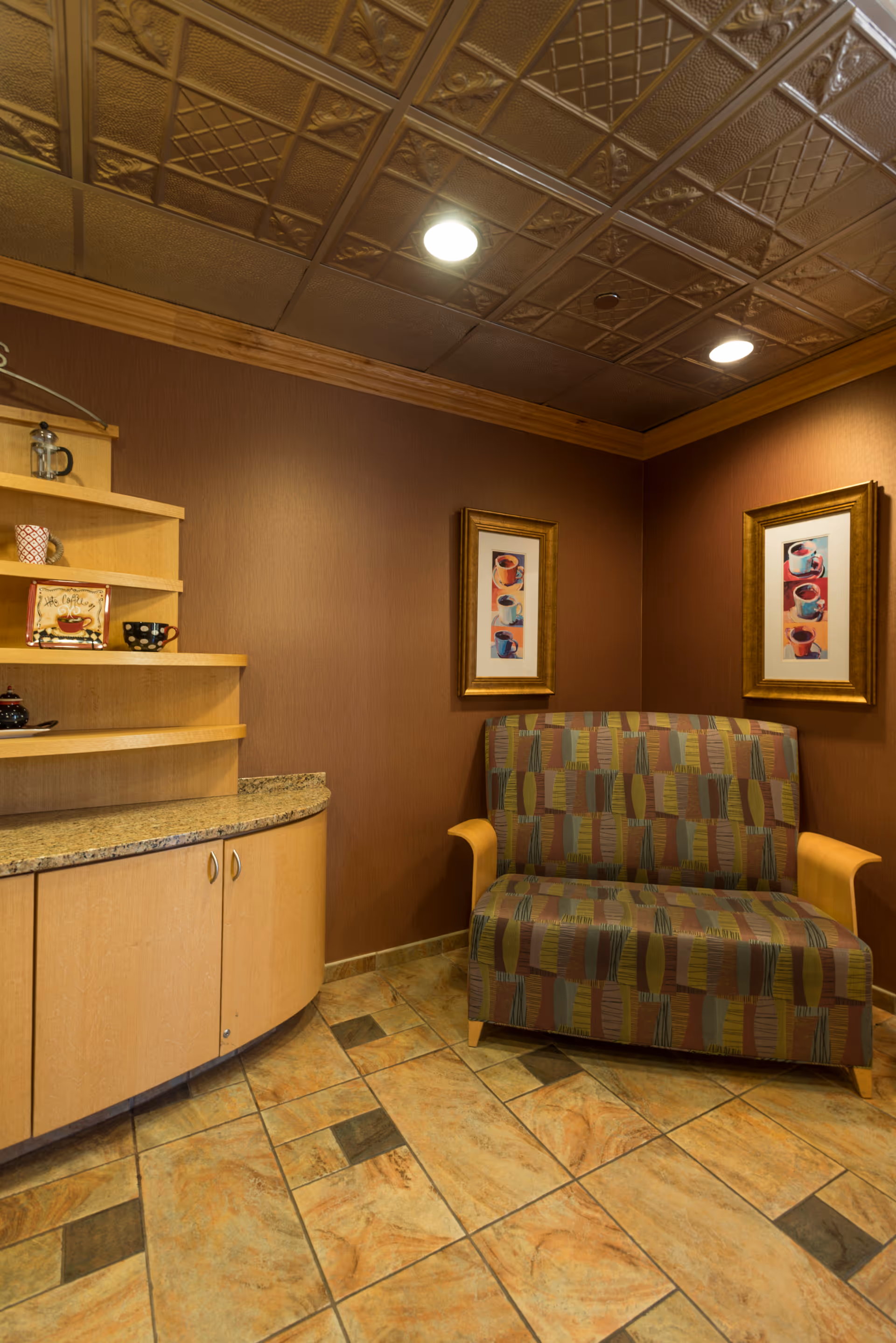 A small seating area with a patterned loveseat against a brown wall, two framed pictures of coffee cups hanging above it, and a curved wooden cabinet with granite countertop and shelves holding decorative cups and a small sign.