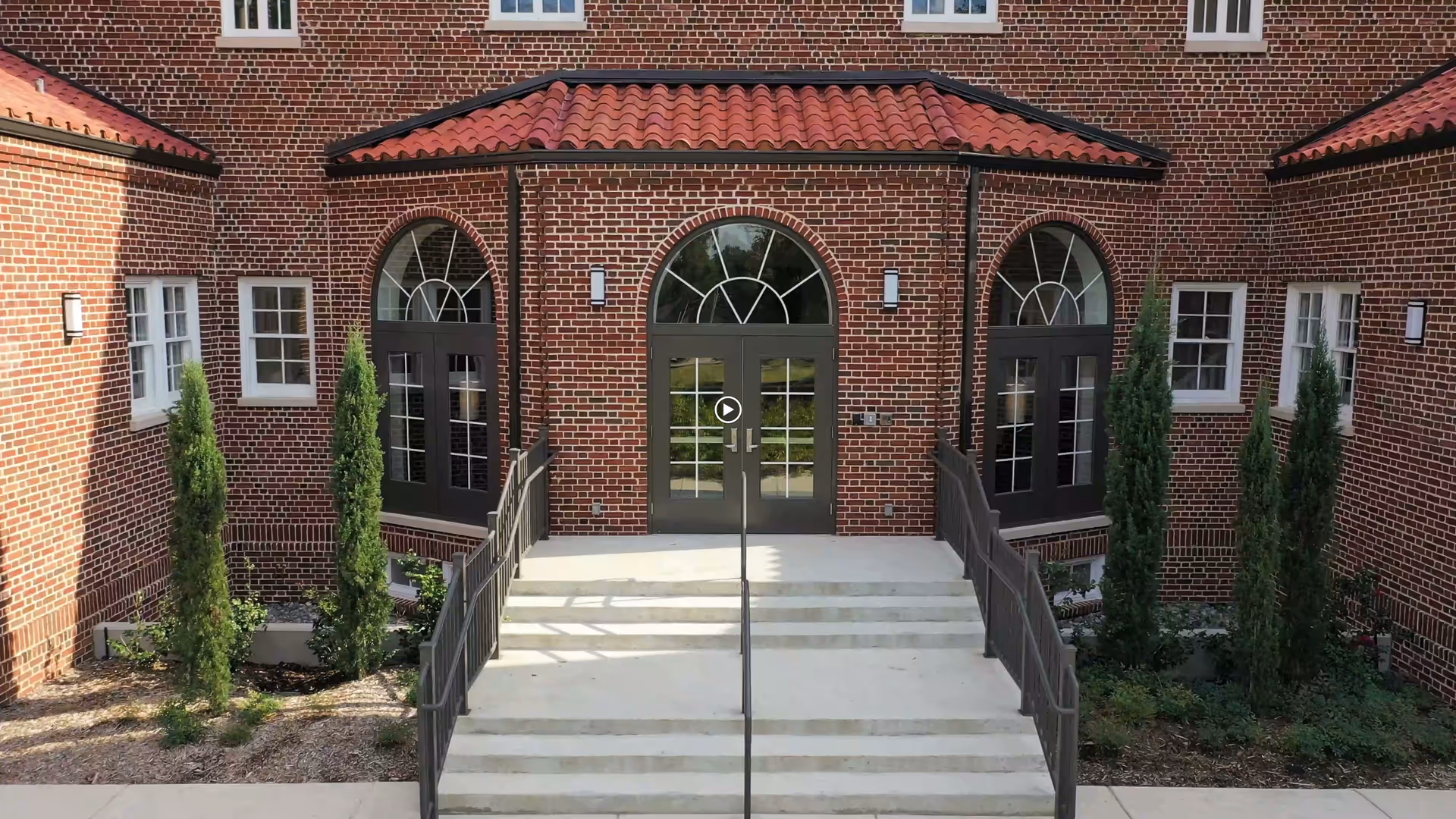 Entrance to a brick building with three arched doorways, each with dark framed glass doors and windows above. There are concrete steps with metal railings leading up to the central door, and small landscaped areas with tall, narrow evergreen shrubs on either side of the steps.