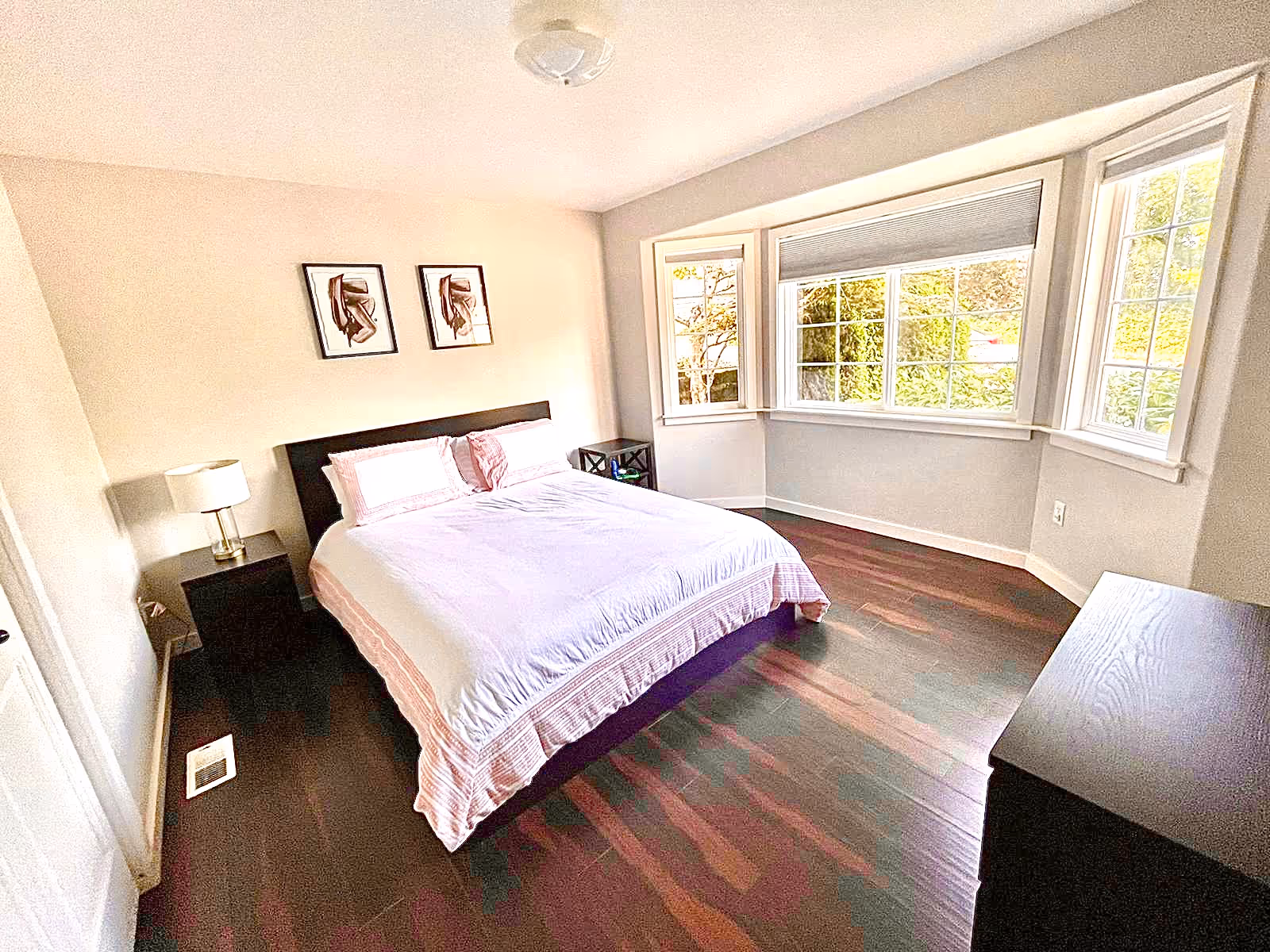Bright bedroom with bed, nightstands, dresser, and large bay windows.