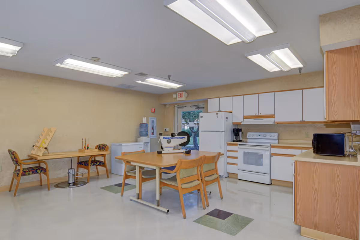 Bright dining area with a central table and adjacent kitchen in a senior facility.