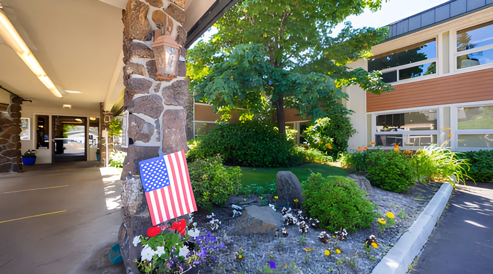 Front entrance and landscaped courtyard of South Hills Rehabilitation Center featuring a stone column with an American flag, flowers, and surrounding greenery.