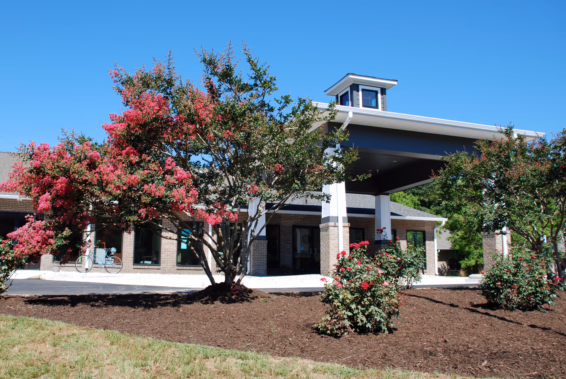 Front entrance of a single-story brick building with a covered porte-cochere, flowering trees, and landscaped beds.