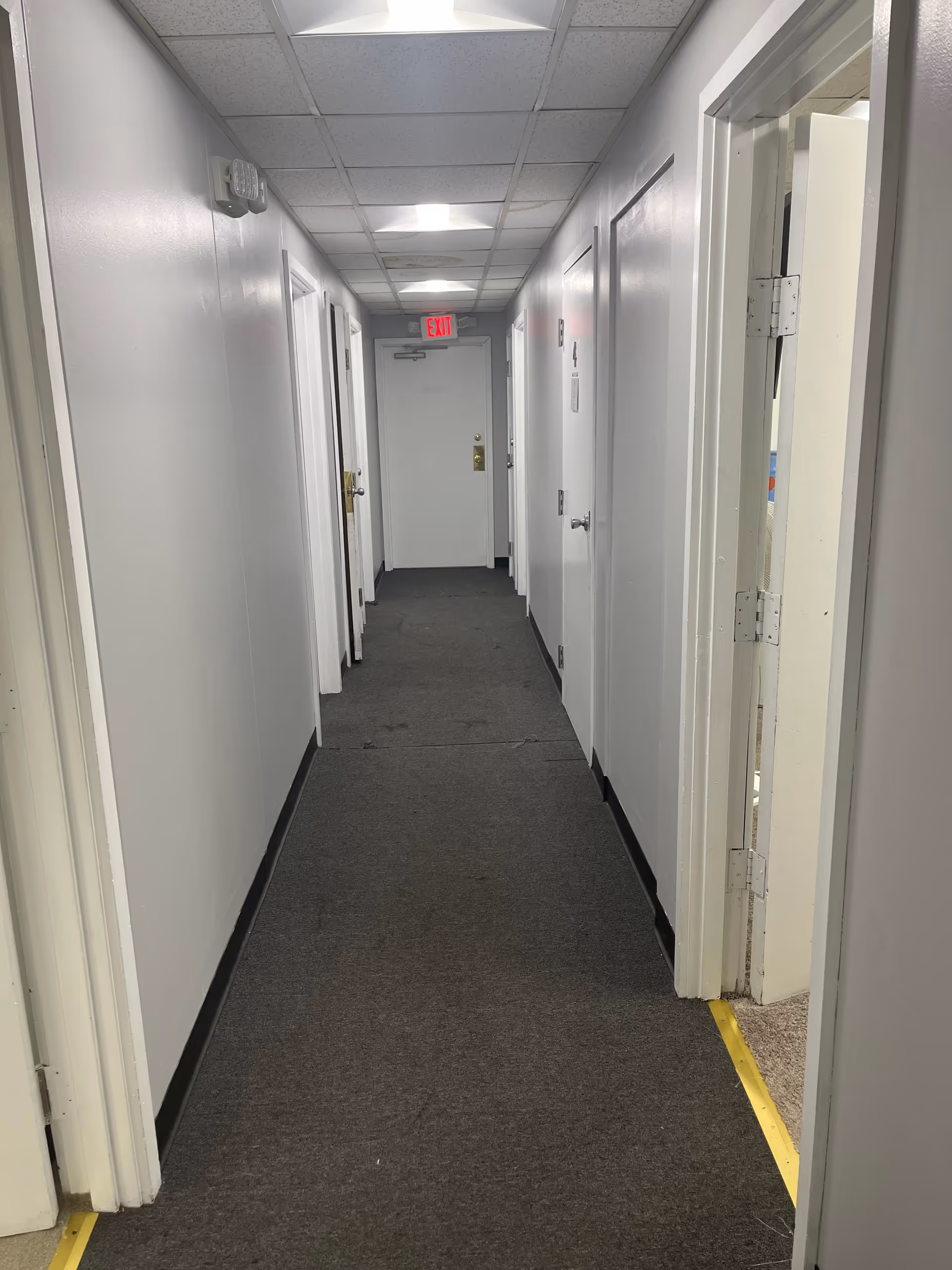 Long narrow corridor with white walls, doors on both sides, EXIT sign overhead.