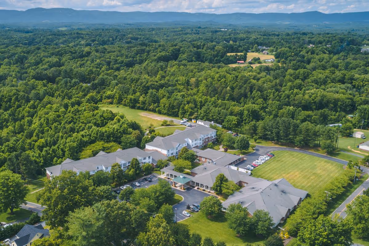 Aerial view of RidgeCrest senior living facility surrounded by dense green trees and open grassy areas, with multiple buildings and parking lots visible under a partly cloudy sky.