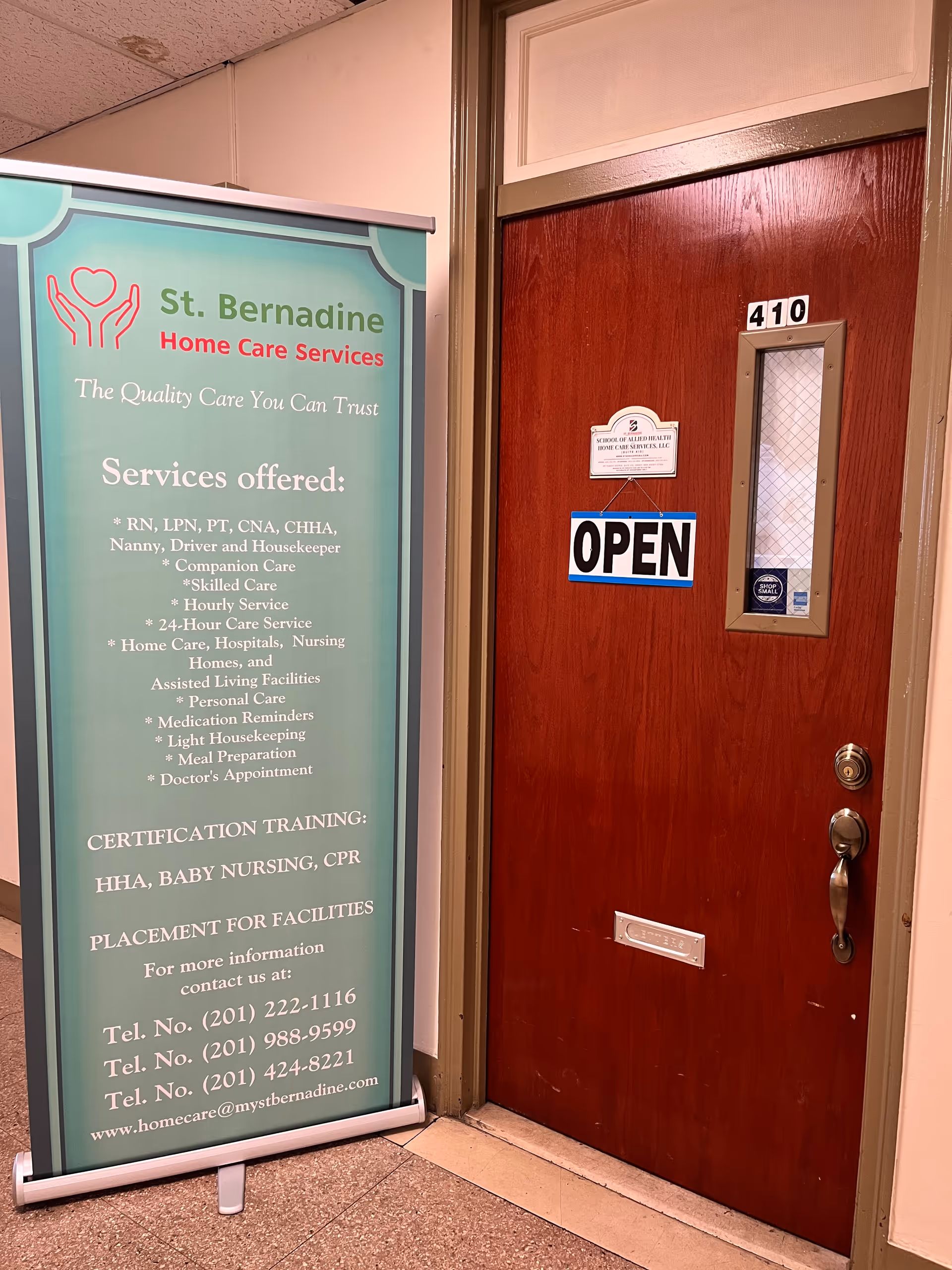Interior hallway with teal St. Bernadine banner beside a wooden door marked 410 reading OPEN.