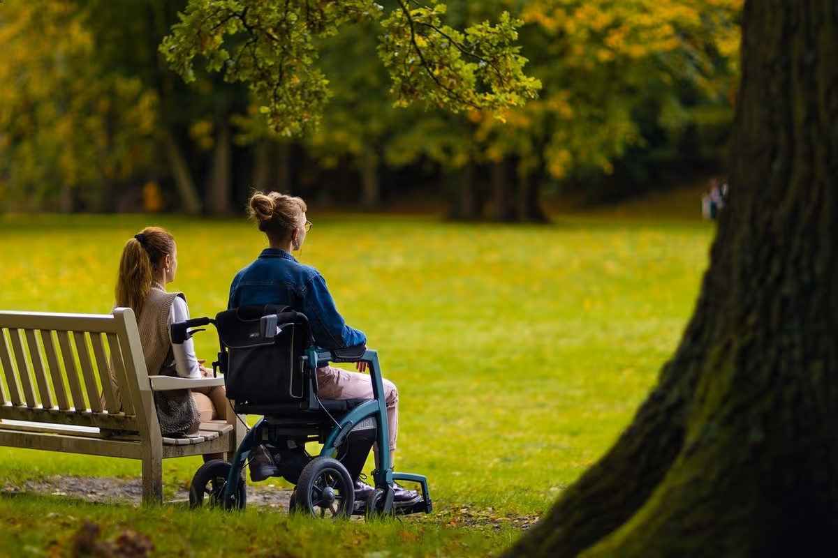 Two people sit on a bench beside a wheelchair in a sunny park.