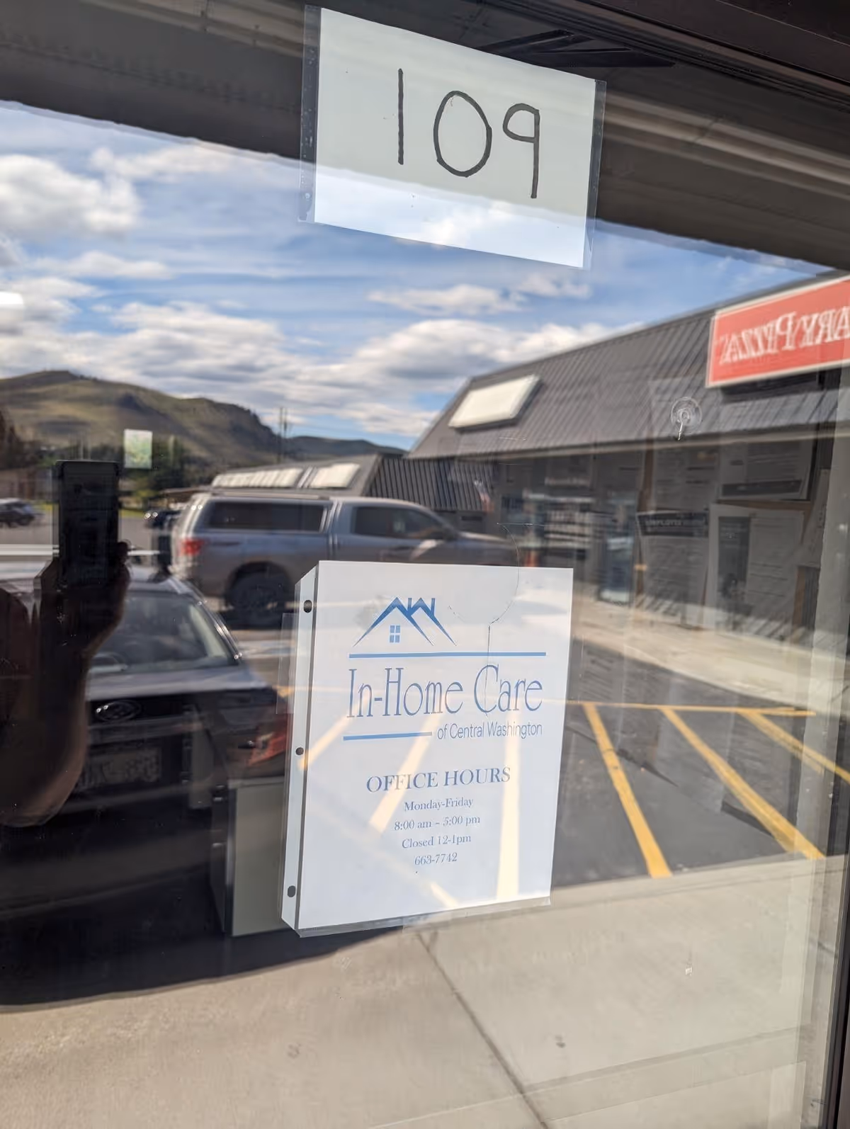 Storefront window shows In-Home Care of Central Washington sign; 109 taped above, reflection shows parking lot.