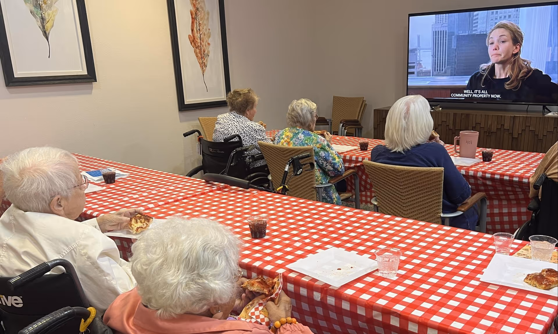 A group of elderly people sitting at tables covered with red and white checkered tablecloths, eating pizza and watching a television mounted on the wall. The room has framed leaf artwork on the walls and the people appear to be in a communal dining or activity area.