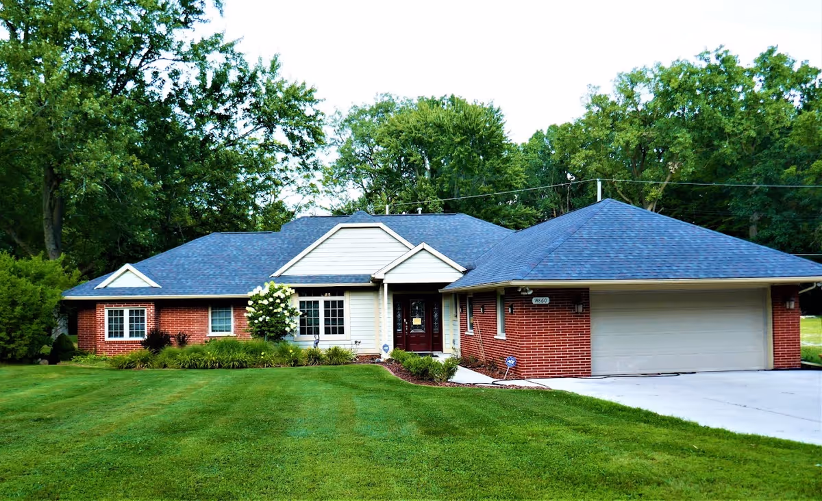 Single-story brick and siding home with an attached two-car garage, manicured front lawn, and trees behind it.