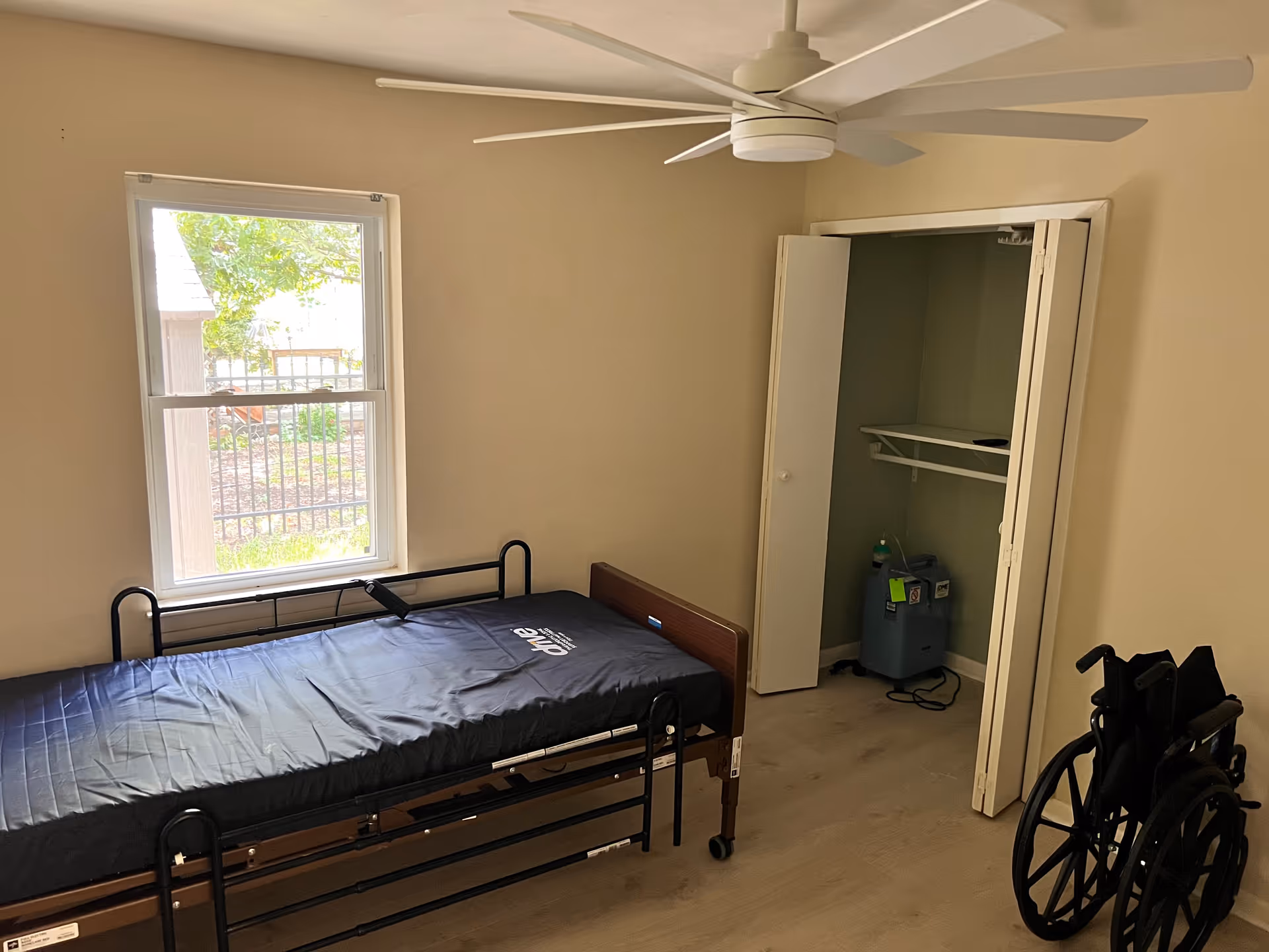 Simple bedroom with a single bed, window, ceiling fan, and a wheelchair nearby.