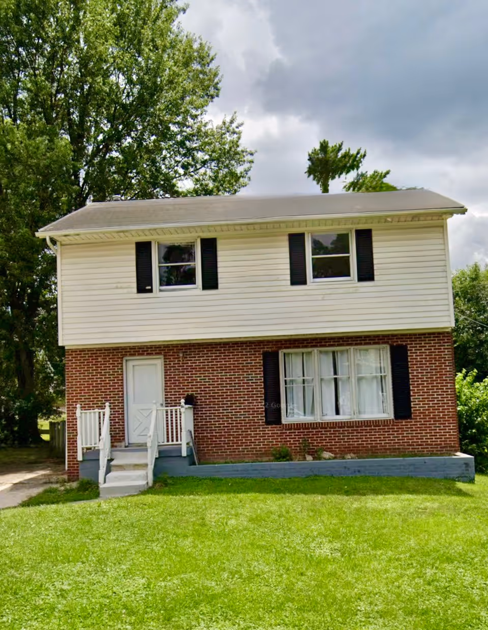 Two-story brick-and-siding building with front steps, door, and large window over a green lawn.