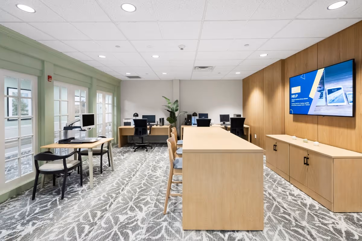 Bright, modern interior common area with desks, computers, a central island, and mounted TV.