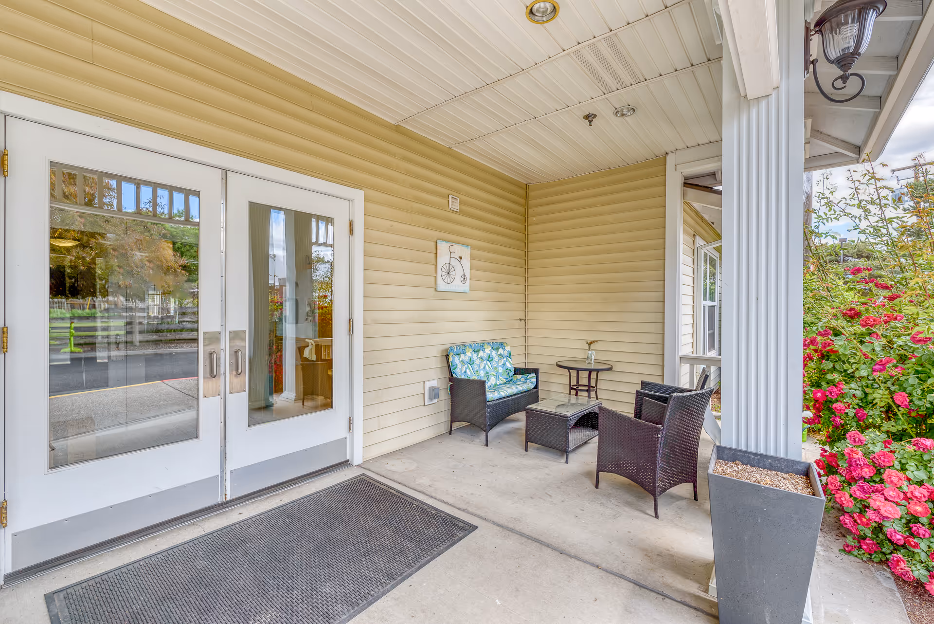 Covered entrance porch with glass double doors, wicker seating and a small table beside flowering shrubs.