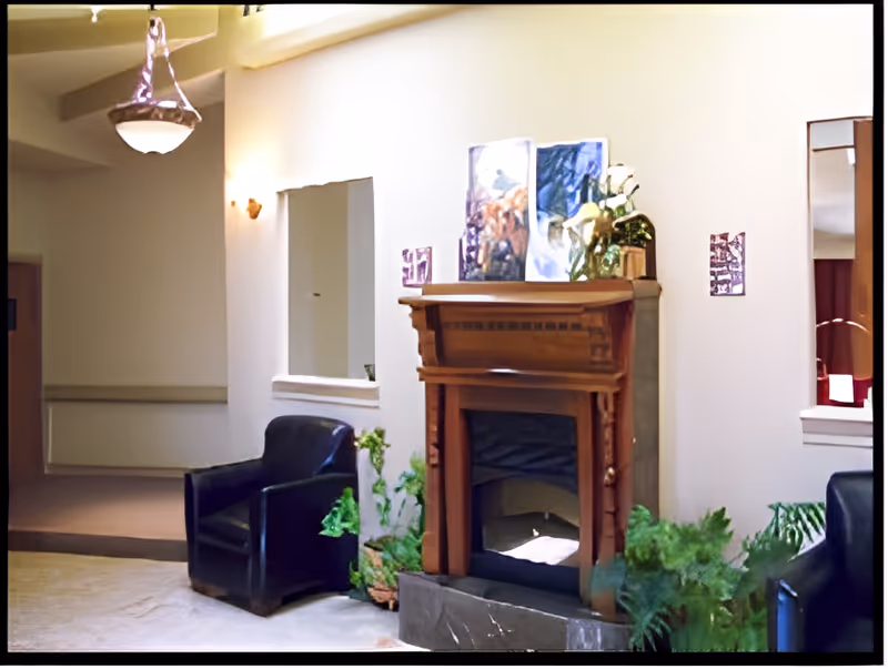 A cozy assisted-living common room featuring a wooden fireplace, leather chairs, potted plants, and wall art.