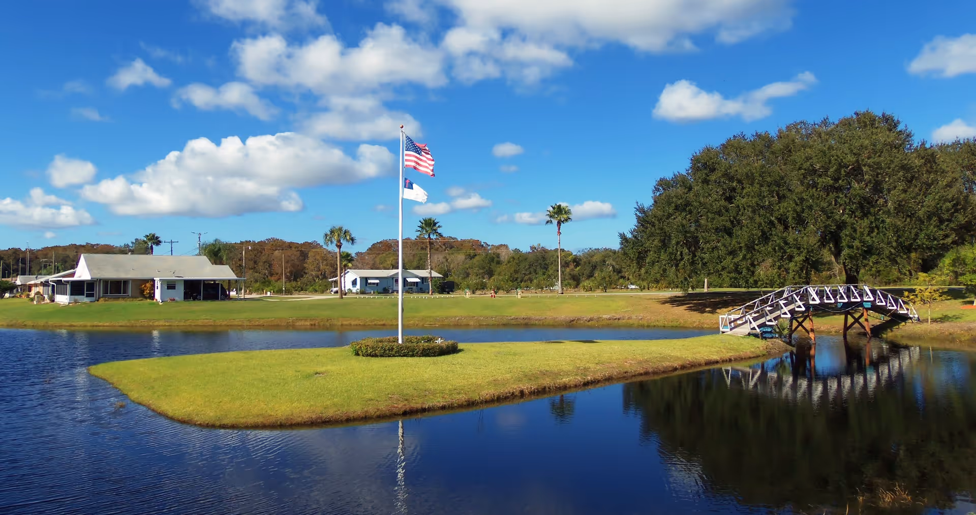 A scenic outdoor view of a small island with an American flag and another flag on a flagpole, surrounded by a pond. There is a white arched wooden bridge connecting the island to the grassy land. In the background, there are trees, a few buildings, and a clear blue sky with some clouds.