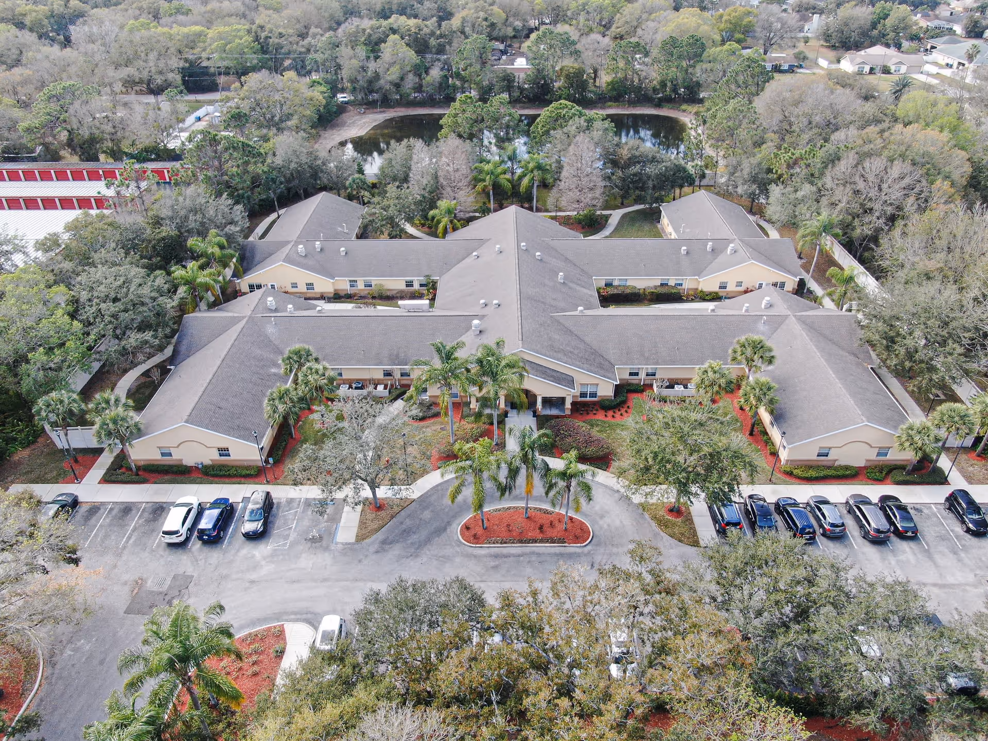 Aerial view of a single-story memory care building with a landscaped circular driveway, palm trees and parked cars in front.